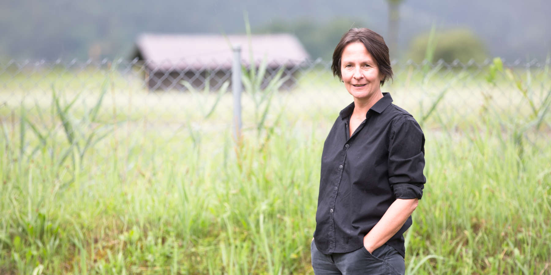 Das Bild zeigt die Expertin für Biodiversität Prof. Almut Arneth wie sie in einem schwarzen Hemd mit den Händen locker in der Hosentasche auf einer grünen Weide steht.