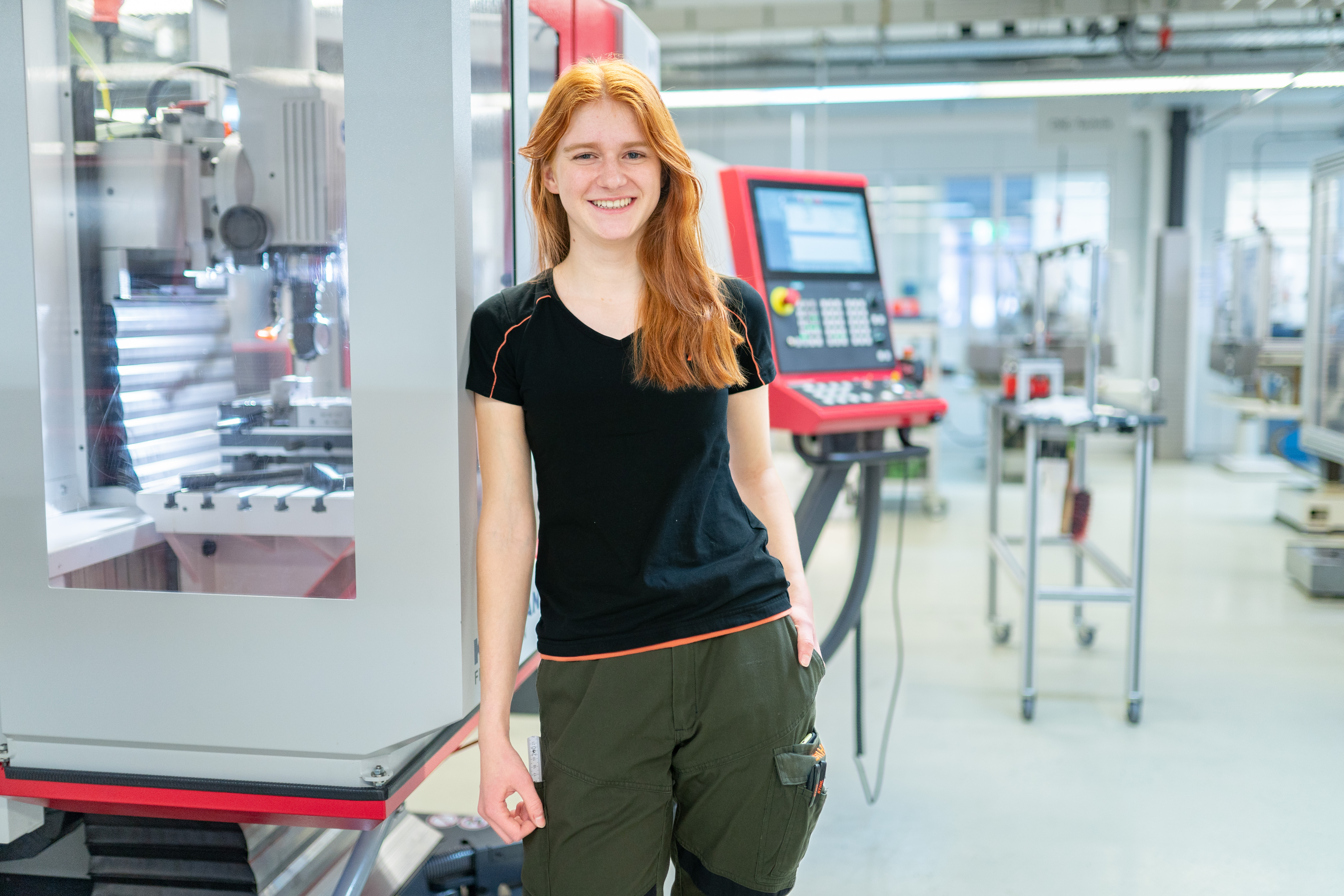 Eine junge Frag mit roten langen Haaren steht in Arbeitskleidung in einer Werkstatt und lächelt in die Kamera