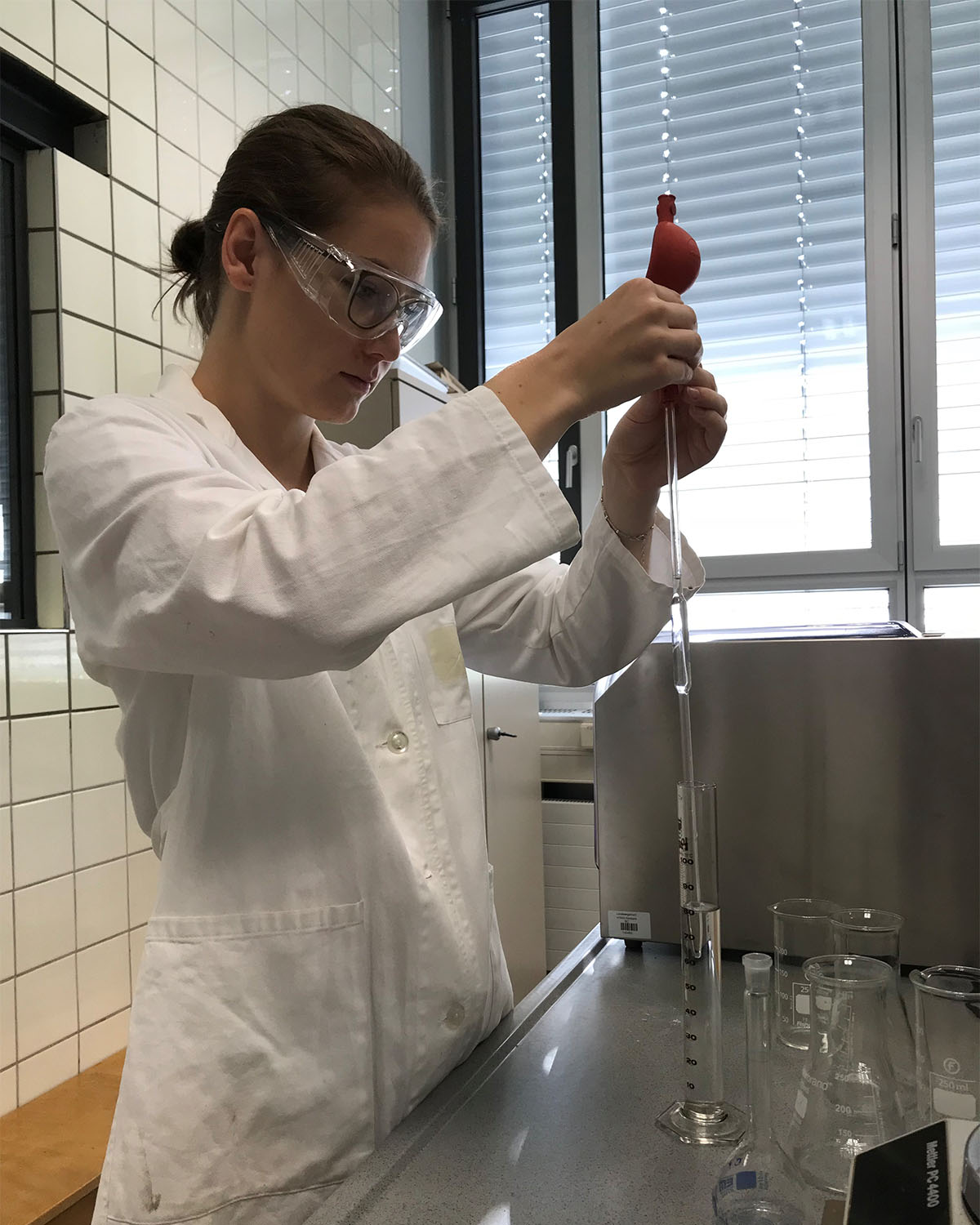 Frau mit weißem Labor Kittel und Schutzbrille macht einen Versuch.