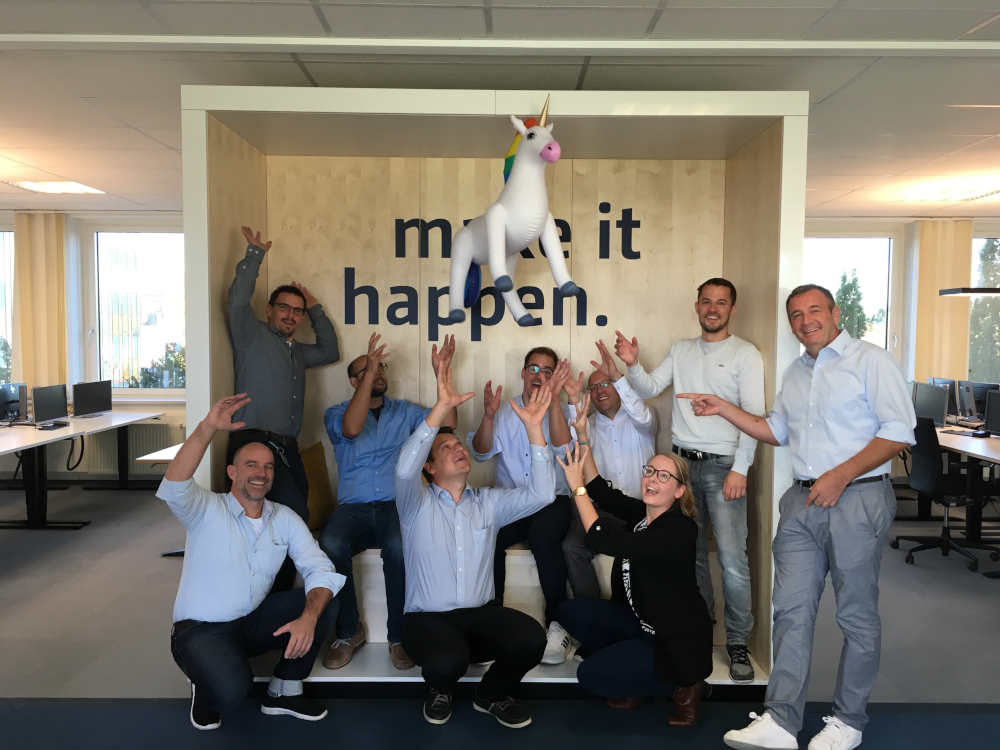 Gruppenbild im Büro von Männern und einer Frau, die die Hände zu einem fliegenden Einhorn Luftballon strecken.