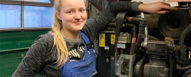 Junge Frau in blauem Arbeitsoutfit steht vor einem Traktorenmotor.