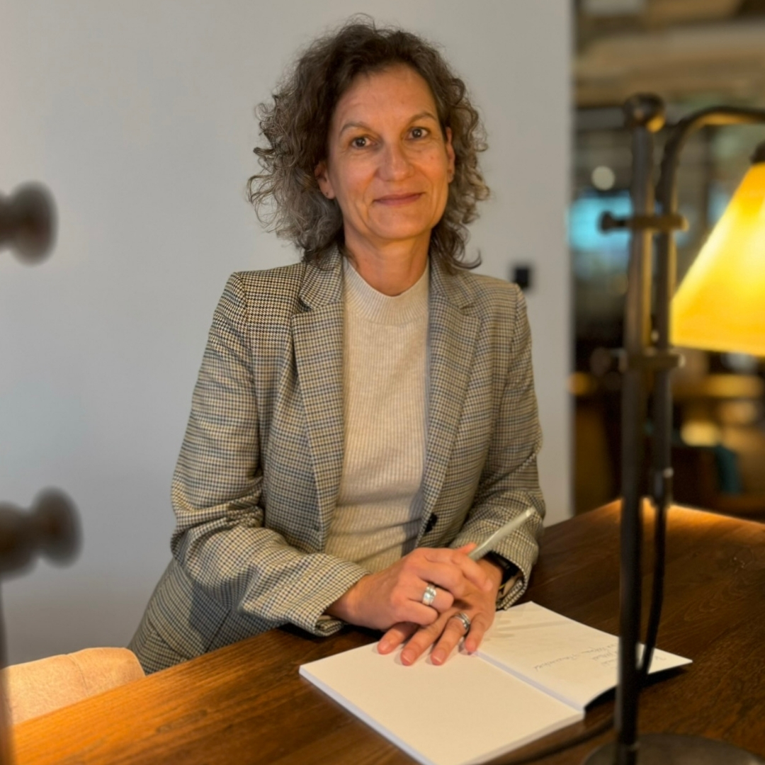 Eine Frau mit schwarz-grauen Locken sitzt an einem Schreibtisch und schaut in die Kamera
