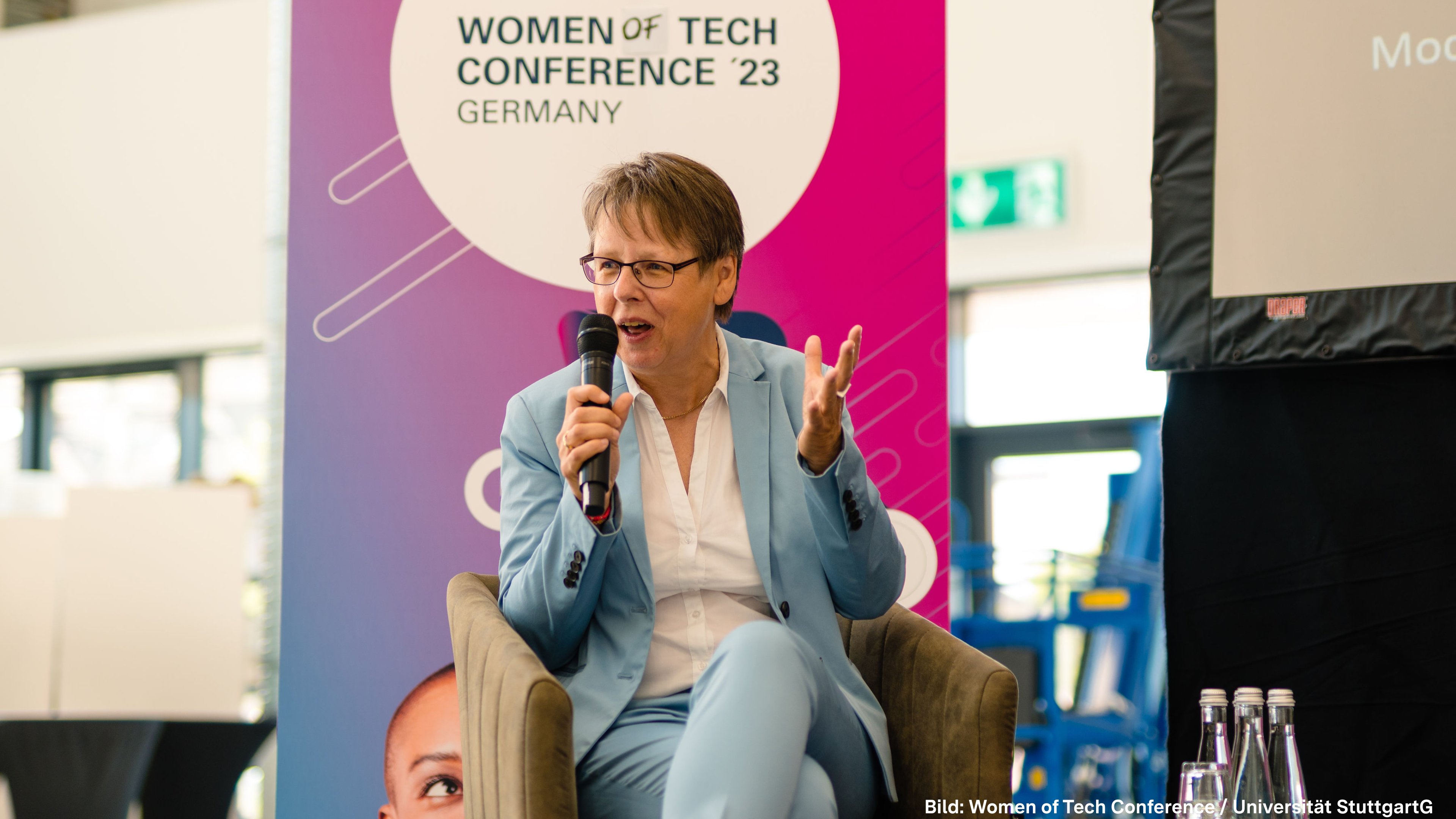 Eine Frau mit kurzen grauen Haaren sitzt auf einer Bühne uns spricht in ein Mikrofon.