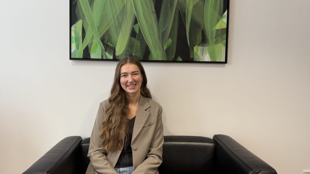 FullSizeRender Eine junge Frau mit grauem Blazer und langen braunen Haaren, sitzt auf einer Couch in einem Büro.